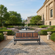 A modern outdoor bench with a dark gray metal frame and brown wood-look seat slats, positioned on a stone paver pathway in a beautifully landscaped garden courtyard with a fountain, hedges, and a classical building in the background.