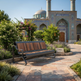 A modern outdoor bench with a dark metal frame and brown wooden slats sits on a stone pathway in a landscaped garden, with a tiled domed mosque in the background.