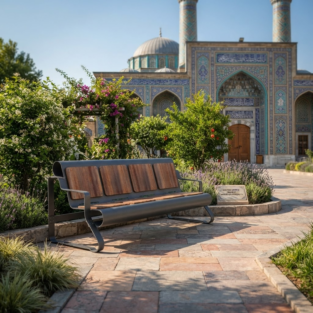 A modern outdoor bench with a dark metal frame and brown wooden slats sits on a stone pathway in a landscaped garden, with a tiled domed mosque in the background.