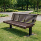 A modern slatted park bench made of dark brown composite material with black metal legs, situated on a green grassy lawn in a park with trees and a paved path in the background.