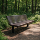 A dark brown slatted outdoor park bench placed on a dirt path surrounded by a dense green forest.