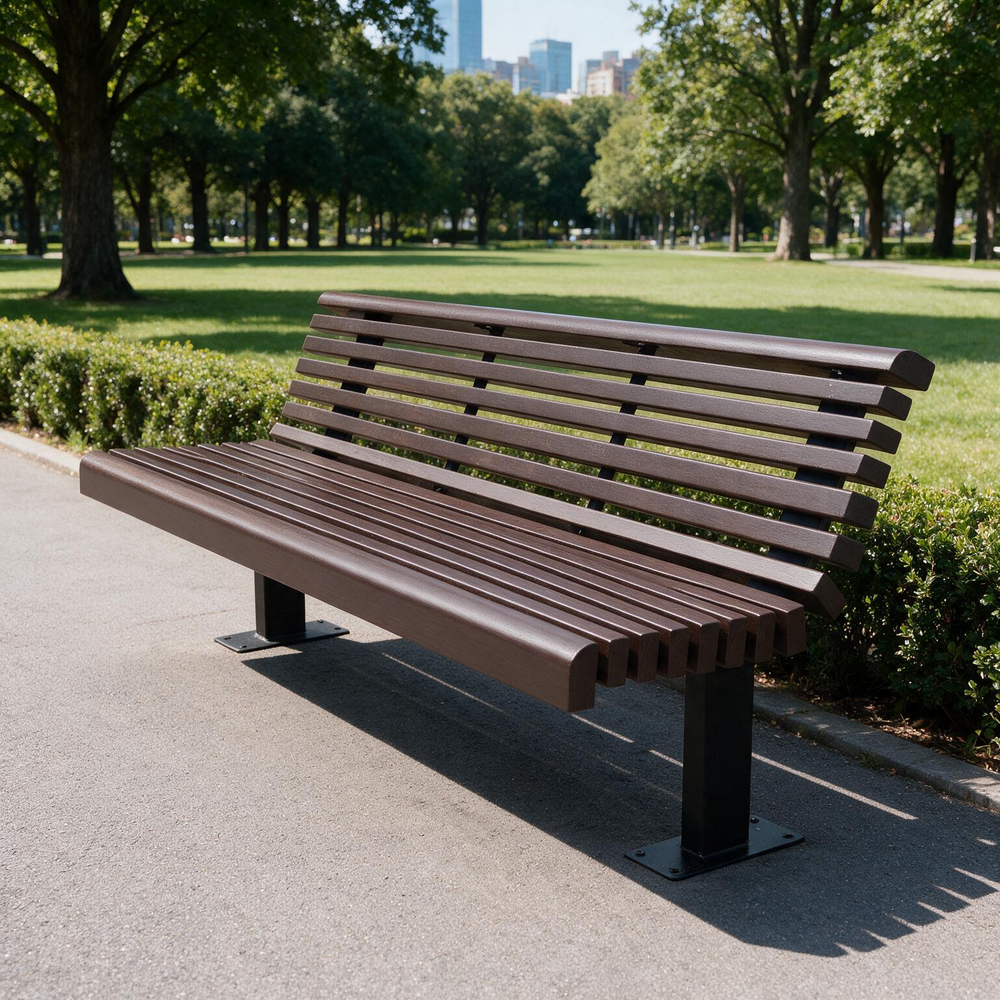 A modern outdoor park bench with brown slatted seating and backrests supported by black metal posts, situated on a paved path in a lush green park with trees and a distant city skyline.