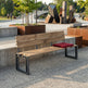 A modern outdoor wooden bench with a slatted back and seat, supported by black metal frame legs, set in a park-like plaza with concrete planters and sculptures in the background. A red cushion rests on the right side of the bench.