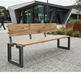 A modern outdoor bench with light natural wood slats and a black metal frame, situated on a paved plaza in front of a contemporary glass building.