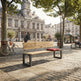 A modern wooden park bench with black metal legs in an outdoor city square, with a red cushion on the seat. The background features historic stone buildings, trees, a bicycle, and people sitting and walking.