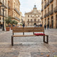 A wooden park bench with dark metal legs sits on a cobblestone plaza in a historic city square, with a red cushion resting on the right side. People walk in the background between tan buildings and a clock tower is visible in the distance.