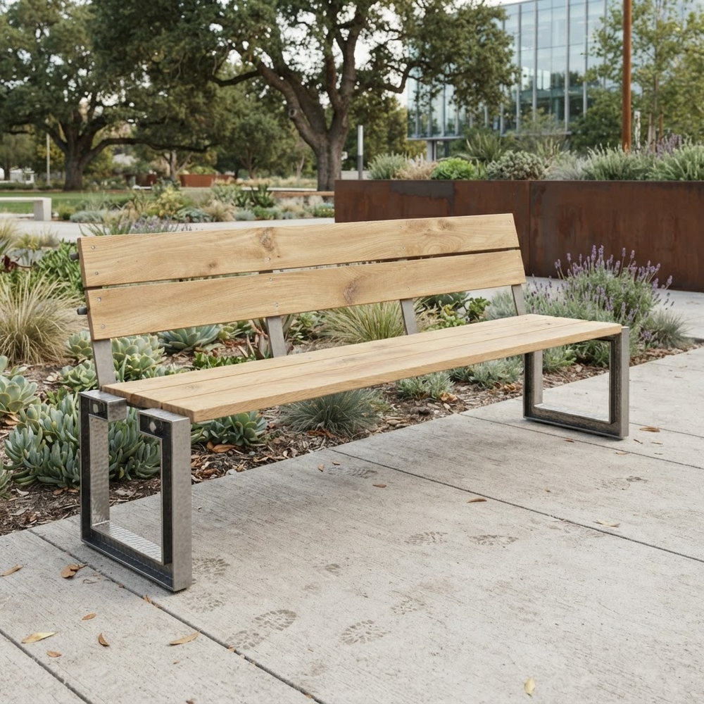 Outdoor park bench with a light natural wood seat and back slats and a dark metal frame, placed on a concrete walkway in a landscaped garden area.