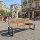 A modern outdoor bench in a European town square, featuring a light oak wood seat and backrest with a dark gray steel frame, and a red cushion on the right side.