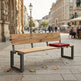 A modern wooden park bench with a red tufted cushion, set on a paved city street with historic stone buildings in the background.