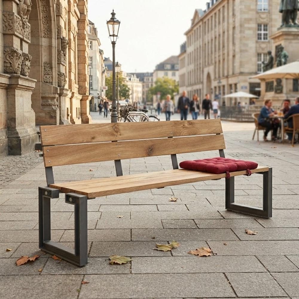 A modern wooden park bench with a red tufted cushion, set on a paved city street with historic stone buildings in the background.