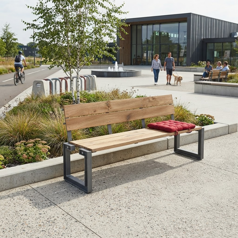 A modern outdoor bench with light natural wood slats and dark metal frame legs, placed on a concrete plaza beside landscaped plants. A red cushion rests on the seat, and people and a cyclist are visible in the background near a contemporary building.