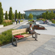 A modern outdoor park bench with a light wood seat and backrest, dark metal frame, and a red cushion, shown in a landscaped plaza with a person sitting on it reading.
