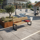 A modern outdoor park bench with a light wood seat and backrest, black metal frame, and a red tufted cushion, placed in a contemporary plaza with people, water features, and greenery in the background.