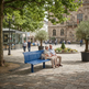 A bright blue outdoor bench in a paved town square, with two elderly people sitting on it. Trees, a small shop, and historic stone buildings are visible in the background.