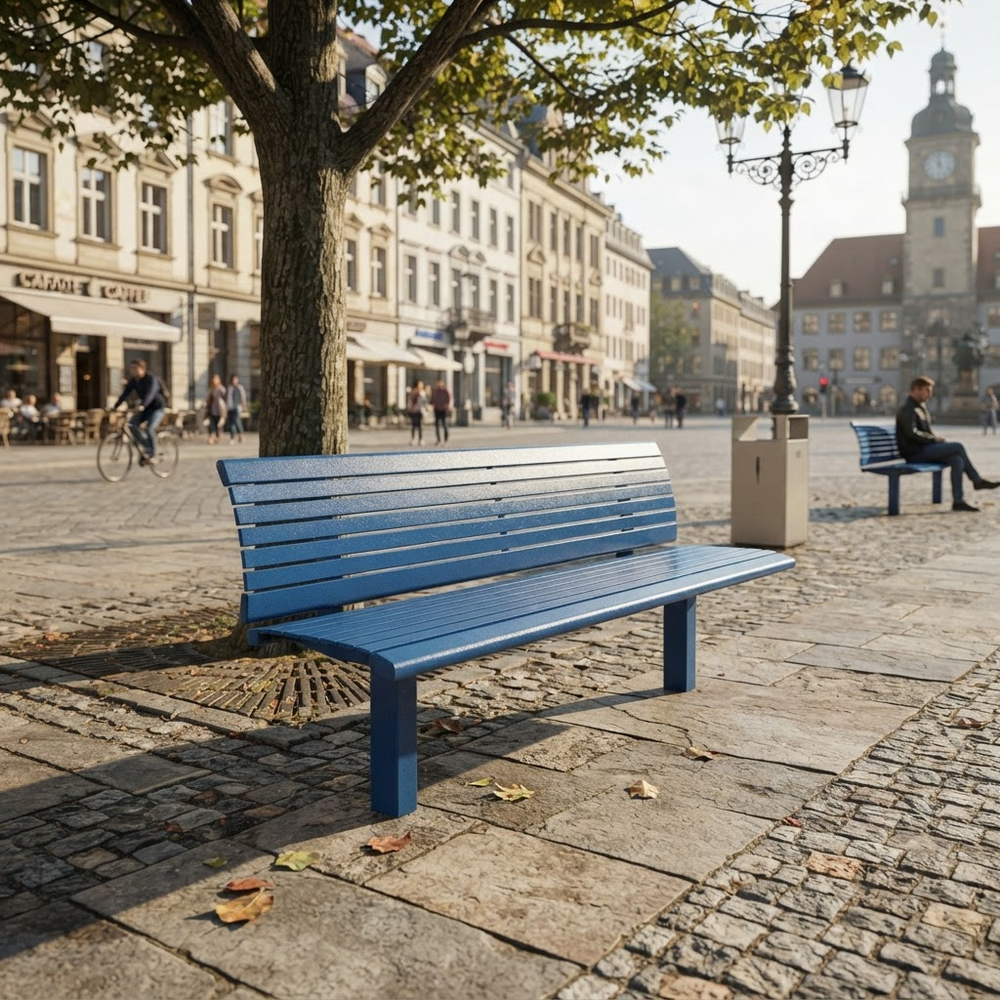 A blue slatted outdoor park bench in a European-style town square, positioned next to a tree on a cobblestone path with buildings and other benches in the background.