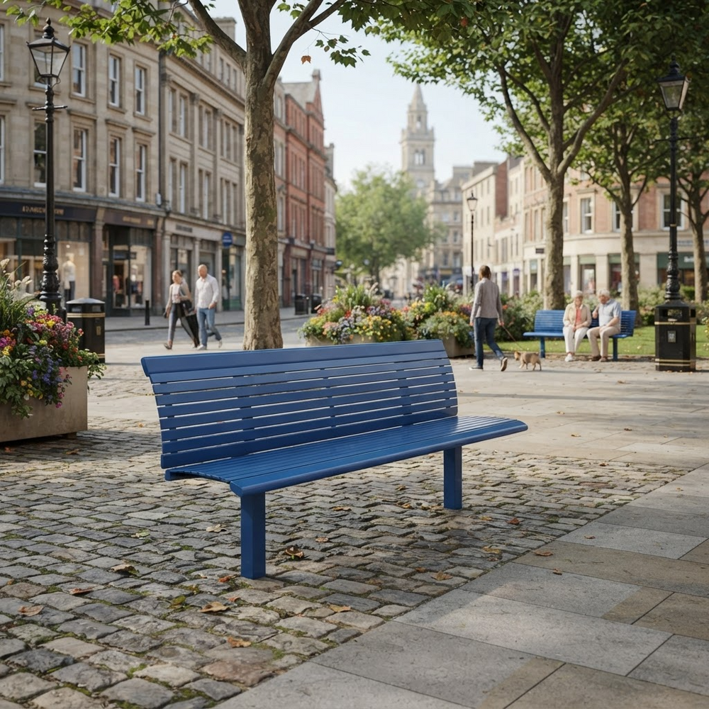 A bright blue slatted outdoor park bench in a cobblestone city plaza, with trees, historic buildings, pedestrians, and flower boxes in the background.