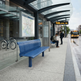A blue slatted public transit bench sits on a city bus stop platform under a glass canopy, with a map display, waiting passengers, a bicycle, and a bus in the background.