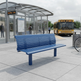 A blue slatted outdoor bench sits on a paved transit station platform, with a yellow bus, station shelter, and a few people visible in the background.