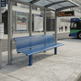 A blue slatted public transit bench placed under a glass bus stop shelter on a city sidewalk, with a bus, street, and station signage visible in the background.