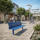 A bright blue outdoor bench in a modern paved shopping plaza with storefronts, trees, pedestrians, and streetlights. The scene appears to be a real environment rather than a clean studio product shot, so this fits Lifestyle rather than Product/Packaging.