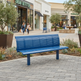 A bright blue slatted outdoor bench placed on a paved plaza in a shopping center, with storefronts, trees, and a few pedestrians visible in the background.