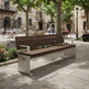 A modern outdoor park bench with dark brown wooden slats and brushed silver metal supports, situated in a sunny stone plaza with trees, historic buildings, and people sitting in the background.