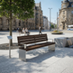 A modern outdoor bench with dark brown wooden slats and brushed silver metal supports, situated in a paved urban plaza with trees, historic buildings, and a few seated people in the background.