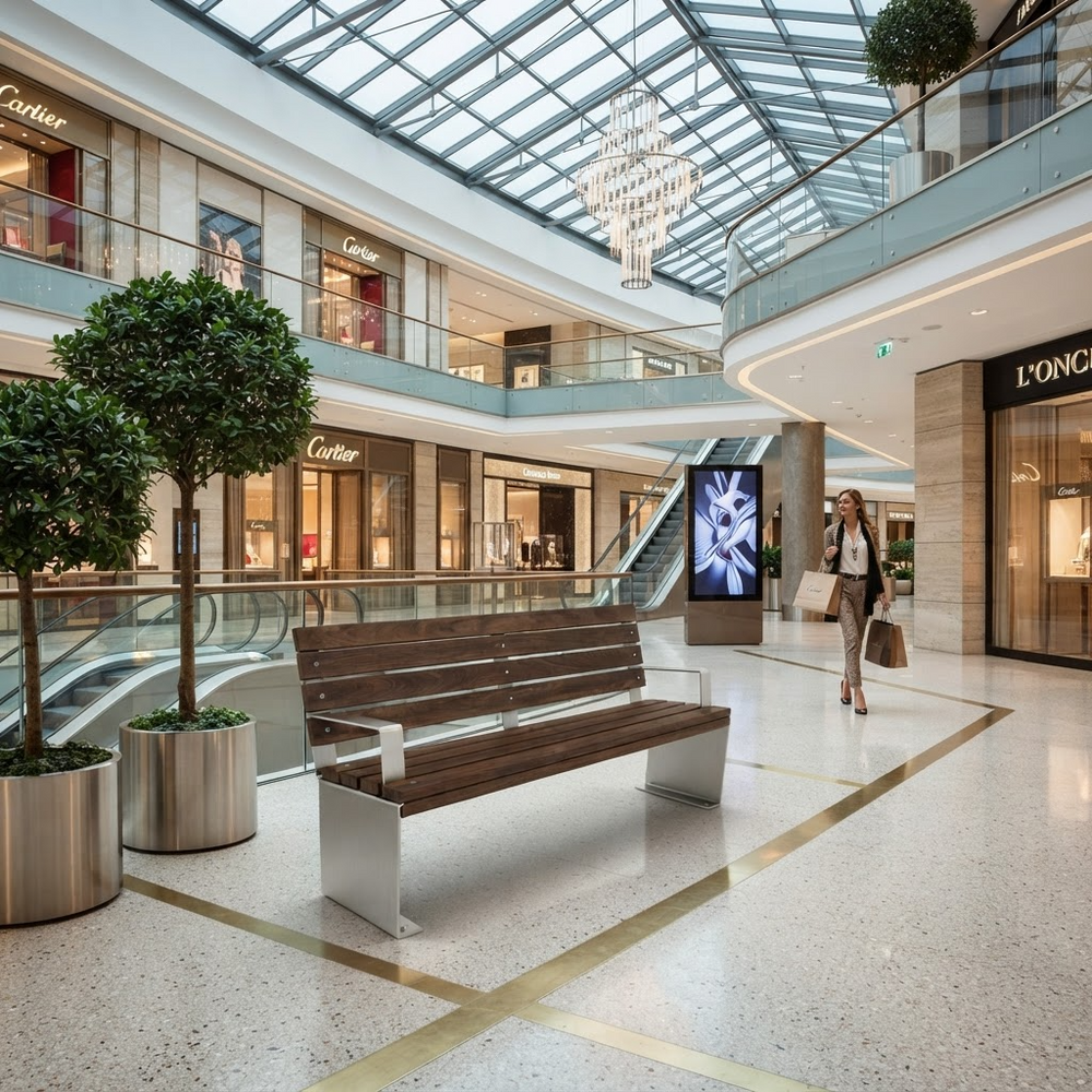 A modern wooden bench with a brushed silver metal base sits in a spacious, upscale shopping mall atrium with a glass ceiling, escalators, and luxury storefronts like Cartier and L'Or.