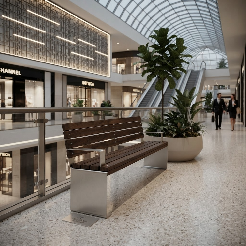 A modern wooden slat bench with a brushed silver metal base sits in a bright, upscale shopping mall corridor with escalators, glass ceiling, and potted plants. Visible storefront signs include Chanel and Bottega Veneta.