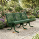 A green metal park bench with a slatted back and seat, placed on a gravel path in a landscaped garden with flowers and trees in the background.