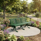 A green slatted park bench sits on a round concrete planter in a landscaped garden with flowers, trees, a lamp post, and a walking path.