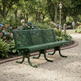 A green metal park bench with curved legs, situated on a gravel path in a lush garden with flowers and trees in the background.
