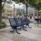 A dark blue slatted outdoor park bench in a city plaza, with trees, pedestrians, seating areas, and modern buildings in the background.