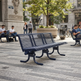 A dark navy blue slatted park bench in a city plaza, with people sitting on other benches and trees in the background.