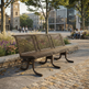 A dark brown wooden park bench with curved metal legs, situated on a paved plaza in a modern urban setting with trees, buildings, and people in the background.