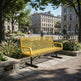 A bright yellow metal bench with a perforated mesh seat and back, situated outdoors on a paved plaza in a park-like urban setting with trees, buildings, and bicycles in the background.