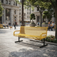 A modern yellow metal mesh bench with black legs in an outdoor urban plaza, with people and buildings blurred in the background.