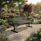 A black metal mesh garden bench placed on a gravel path in a lush, colorful flower garden with trees, a pond, and a gazebo in the background.