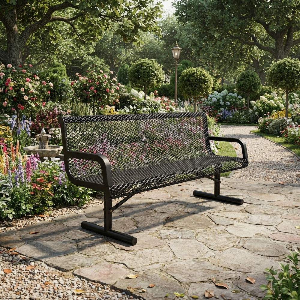 A black metal mesh park bench with armrests, positioned on a stone pathway in a lush, green garden with flowers and trees in the background.