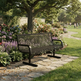 A black metal mesh park bench sits on a stone pathway in a lush, green backyard garden with flowers and a tree in the background.