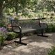 A black metal mesh park bench with curved armrests, situated on a gravel path in a lush, green garden with flowers and trees.