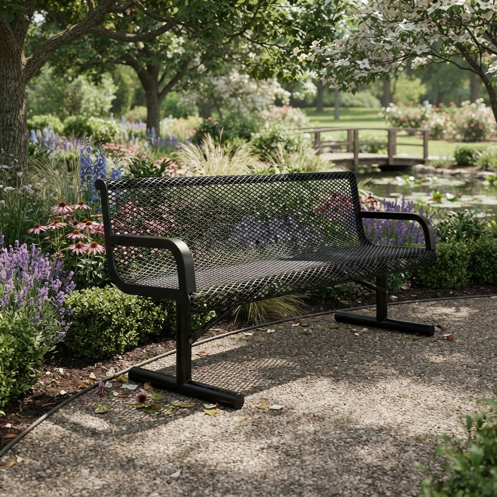 A black metal mesh park bench with curved armrests, situated on a gravel path in a lush, green garden with flowers and trees.