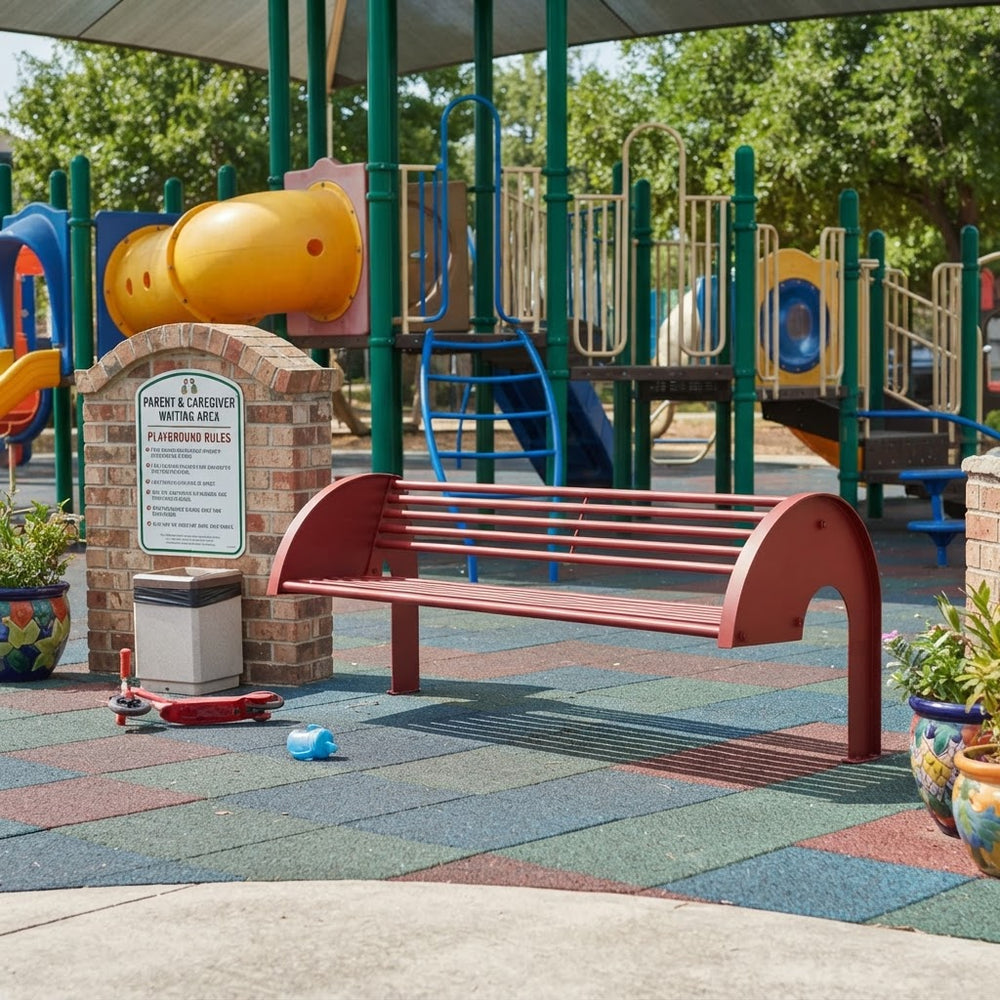 A red metal park bench sits on a colorful rubberized playground floor in front of a brick sign and a large playground structure with green poles and yellow slides.