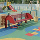 A red metal playground bench with curved backrests and seat slats, placed on a colorful rubberized playground surface with a slide, fence, and toys in the background.