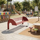 A red metal park bench in a playground setting, with children and adults sitting on benches in the background, trees, and a play structure visible.