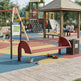 A red metal playground bench with curved ends is shown in a children's playground setting. The bench is positioned on a colorful rubberized safety surface next to a dark brown recycling bin, with playground equipment, a pavilion, and children visible in the background.