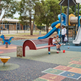 A children's playground scene with a red bench, blue slide, climbing structure, hopscotch markings, and children playing outdoors near trees and a fence.