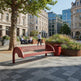 A red curved park bench in an urban plaza with a tree, planters, seated people, and modern buildings in the background.