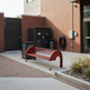 A red outdoor bench placed in a paved courtyard beside a brick building, with recycling bins, a hose reel, and landscaping visible in the background.