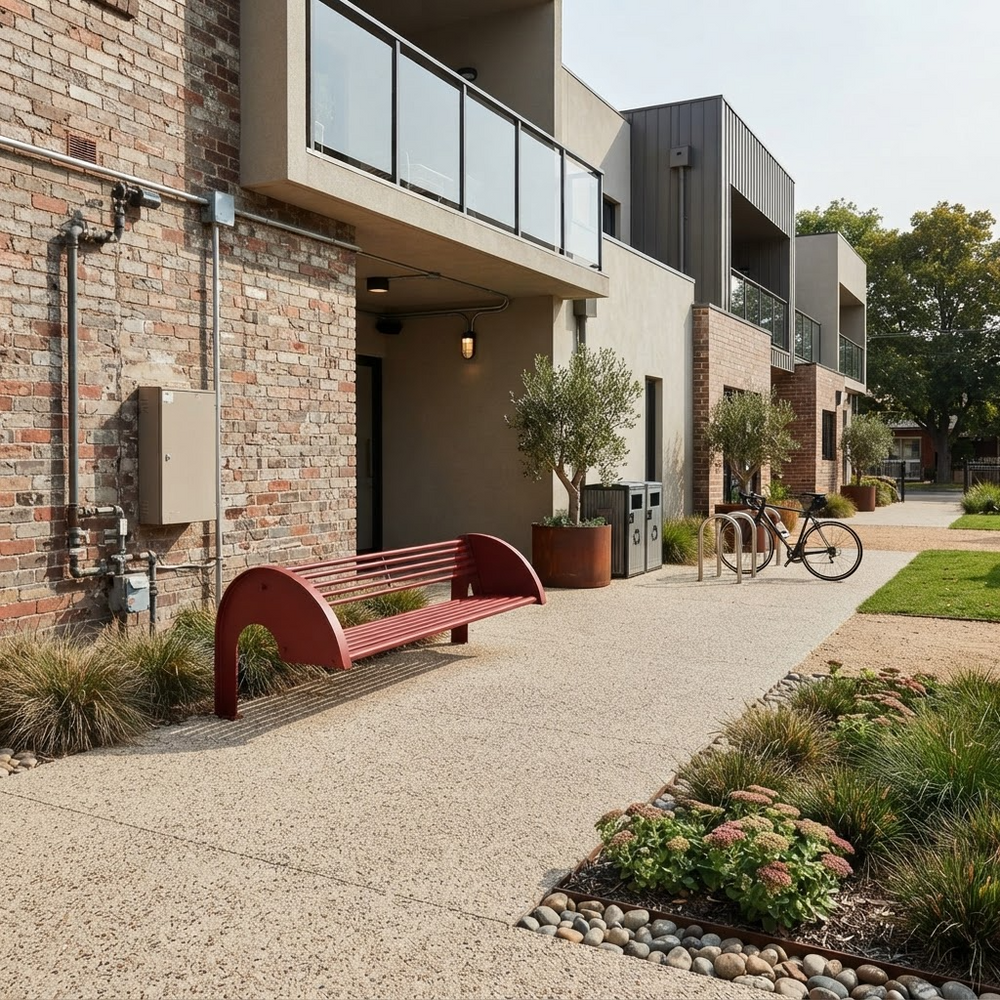 A modern outdoor residential courtyard with a curved red bench in the foreground, a concrete walkway, landscaped planters, and contemporary brick and concrete apartment buildings in the background.